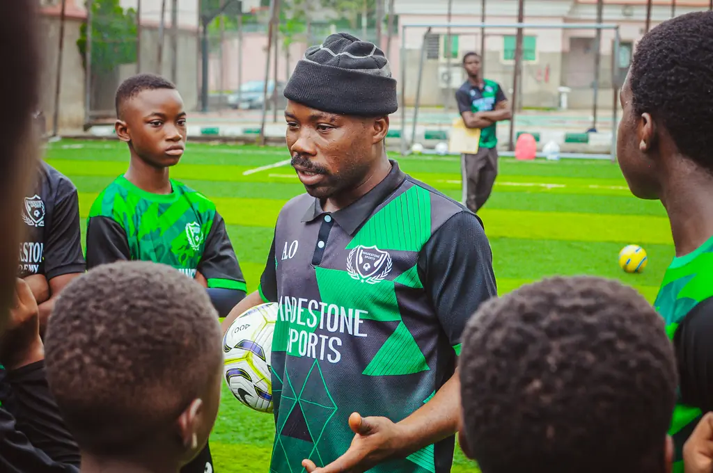 A coach holding a football and speaking to a huddle of young Madestone players